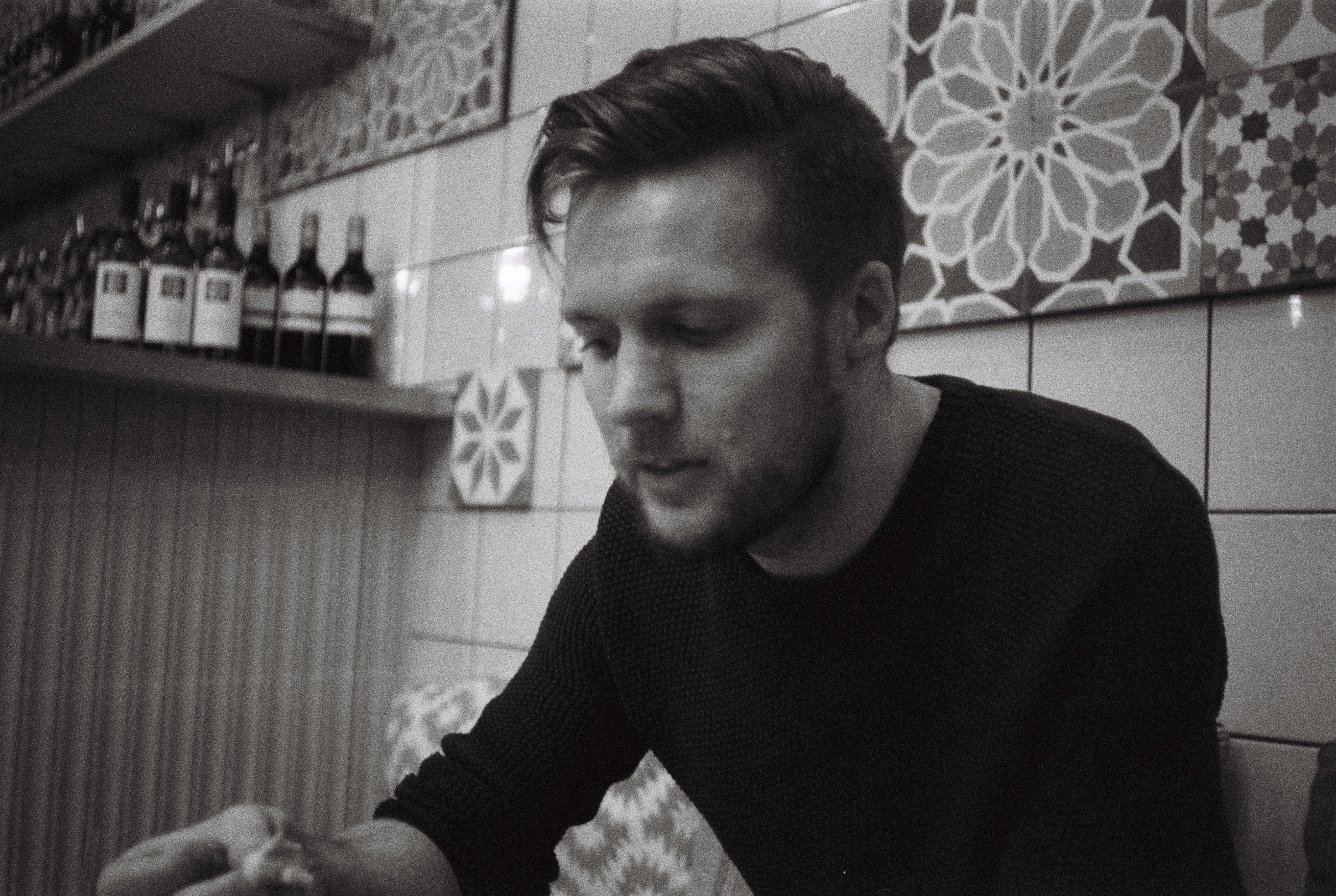 A black and white portrait of coxy (Matt Cox) sitting in front of a tiled wall in a cafe