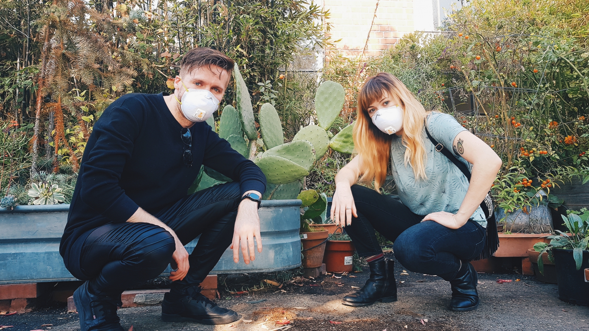 A photo of coxy (left) and Kelly Mason (right) both crouching down infront of some cactus and wearing masks over their mouth