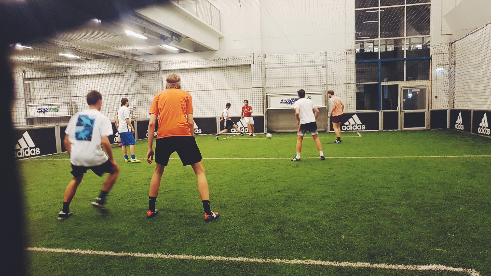 A photo of a small indoor football pitch with a group of boys mid-game, kicking a ball around the surface of the artificial grass.