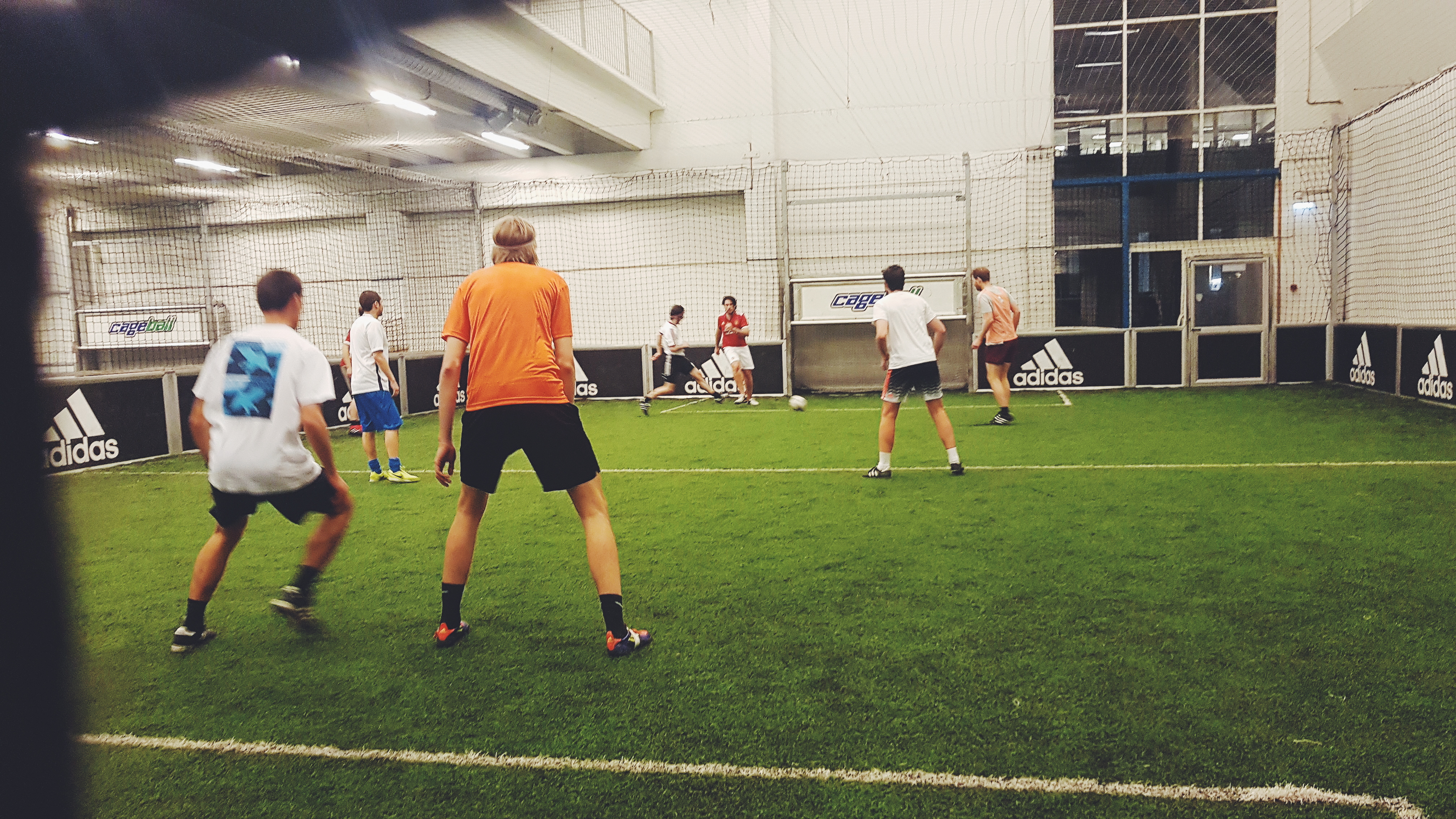 A photo of a small indoor football pitch with a group of boys mid-game, kicking a ball around the surface of the artificial grass.