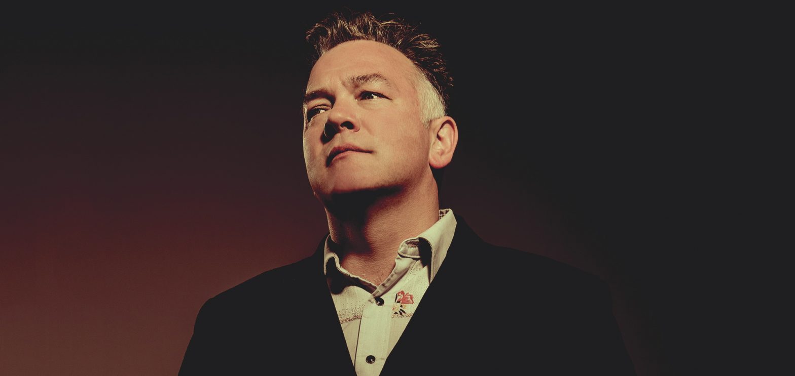 A portrait of stand-up comedian Stewart Lee, wearing a black jacket and looking serious against a dark red background.