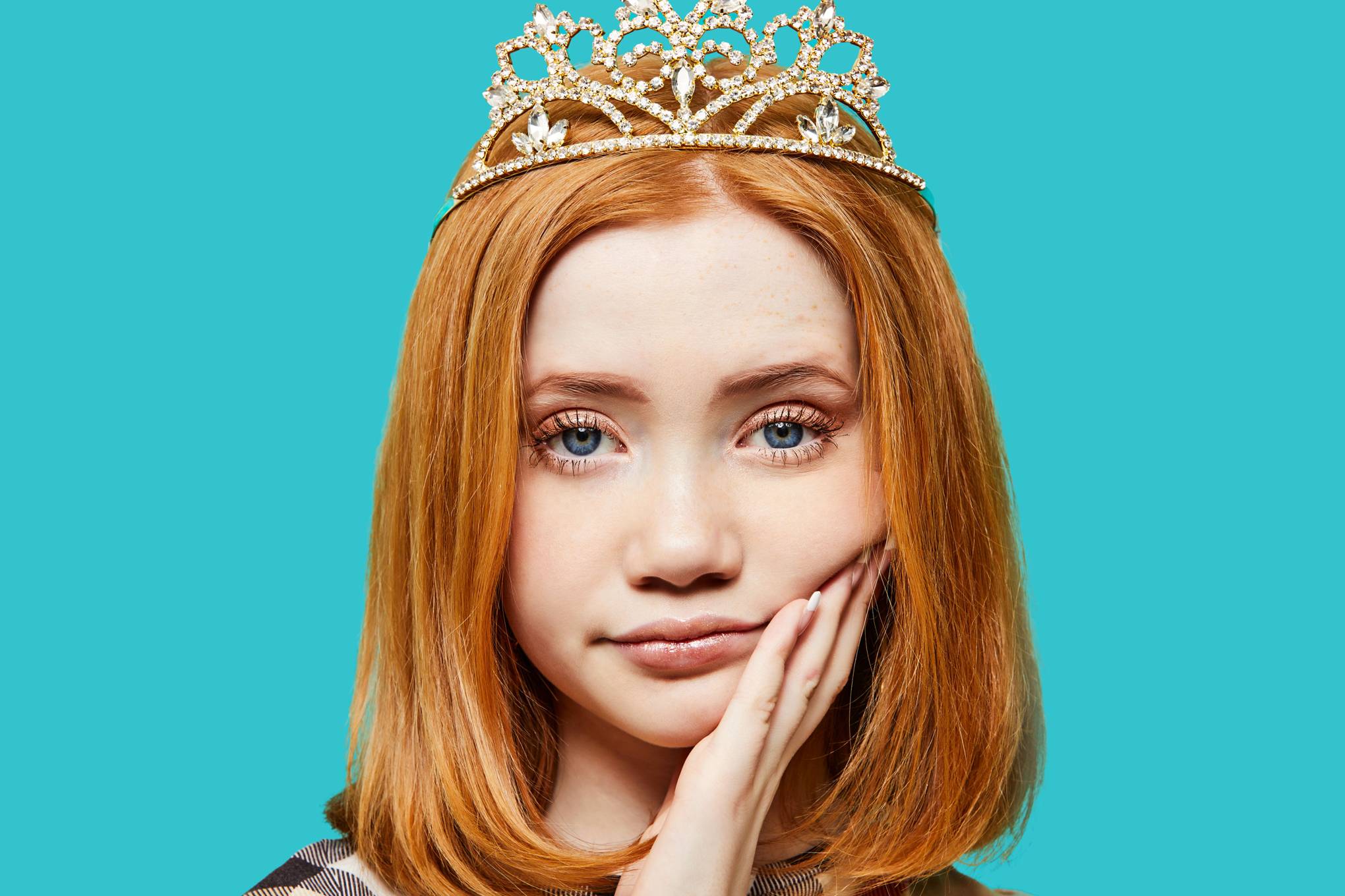 A portrait of a young ginger teenage girl with ginger hear down to her shoulders. She wears a crown on a her head and a non-plussed look on her face as she rests her head in her hand.