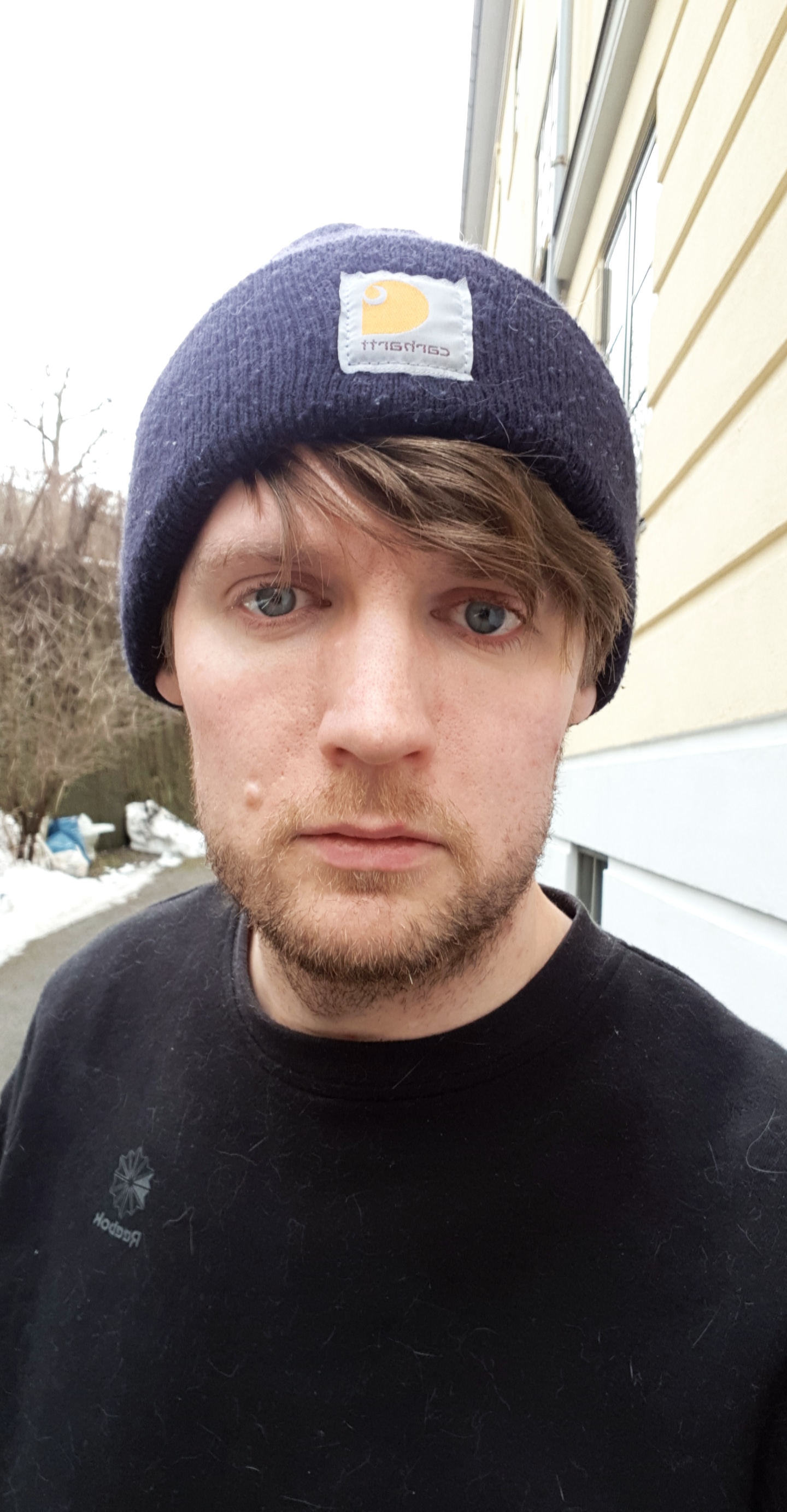A photo of coxy (Matt Cox) looking sraight into the camera, wearing a navy blue beanie hat with a stiched on Carrahatt logo, covering his hear which peaks through underneath, swept across his forehead and slight whisping over his eyes. He is outsite, stood by a beige apartment block.