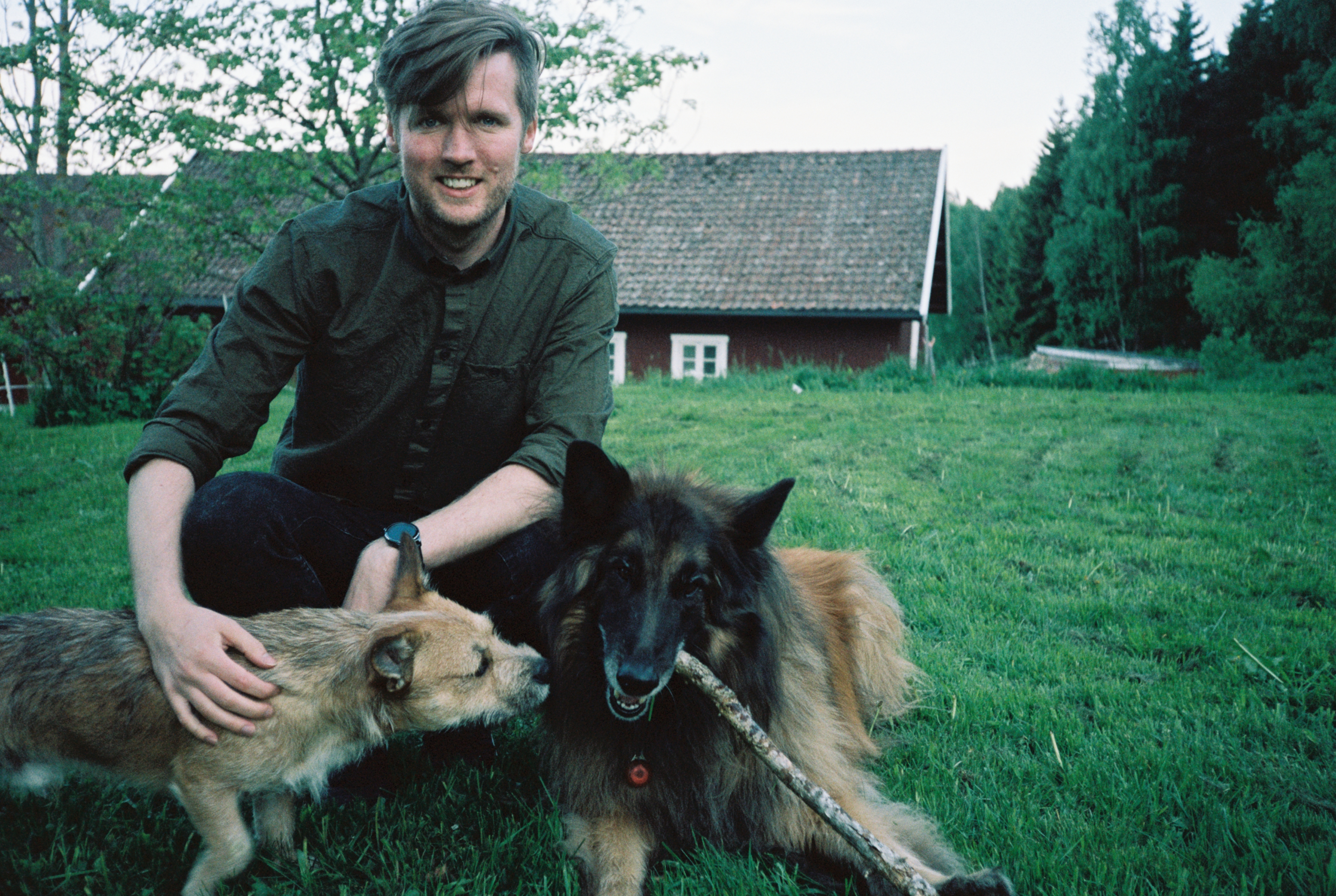 A portrait of coxy (Matt Cox) playing wit two dogs on some grassland in front of a stables.