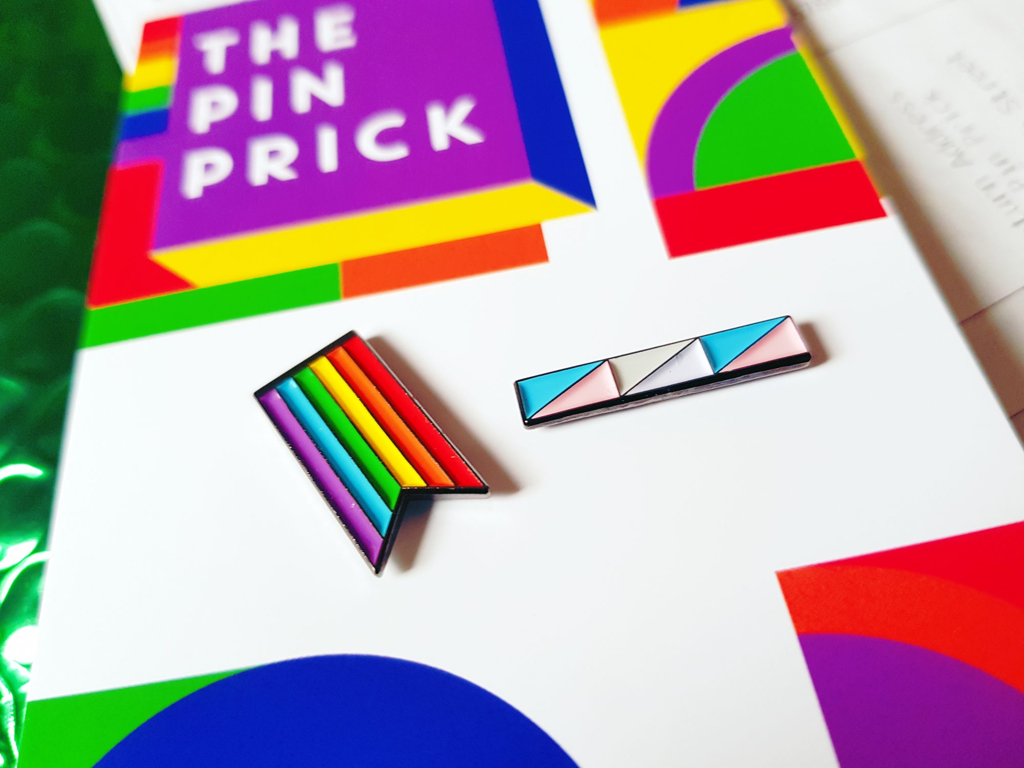 A close up photo of two enamel pin badges. The badge on the left is a rainbow flag, the badge on the right is a long bar with pink, blue, and white shades - representing transgender flag.