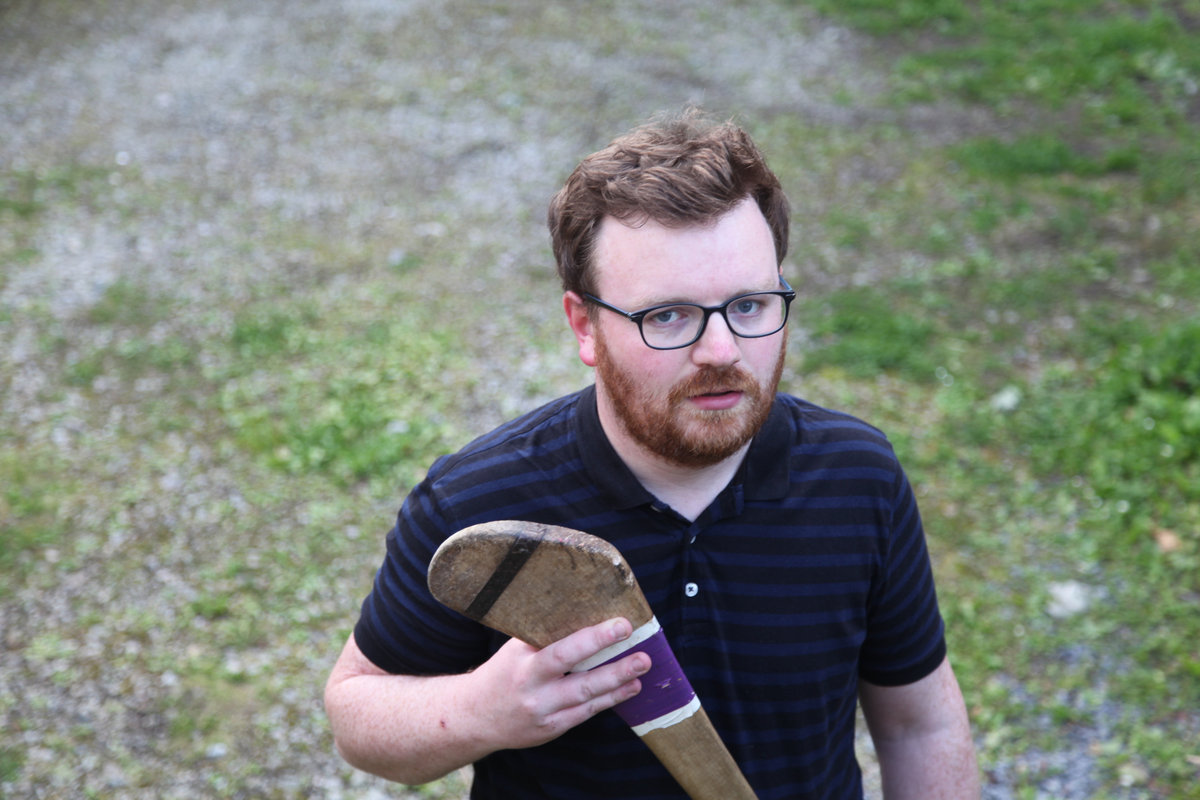 A photo from a slightly raised angle of Brian Kelly, singer and front-person of the band So Cow. He holds a hurl - the wooden stick they use in the Irish sport called hurling.