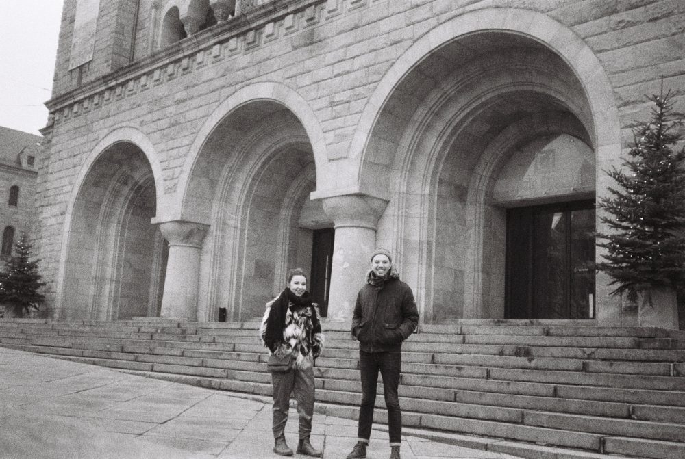 A black and white photo of Ine Vestengen-Cox and Ben J Wood stood together outside a building in Poland