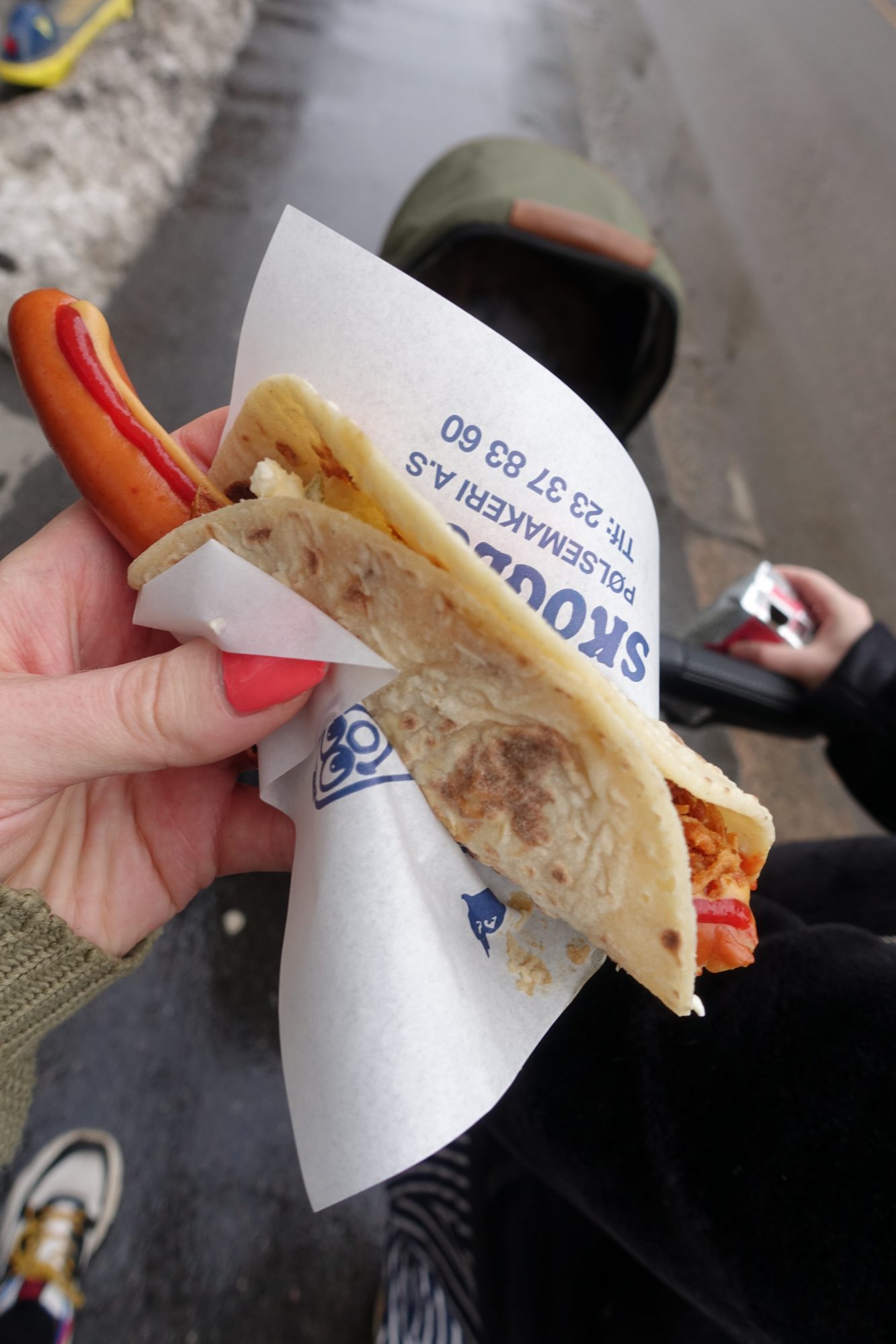 A hand holding a hot dog, covered in tomato ketchup, mustard, potato salad and dried onion - all wrapped in a lompe (norwegian potato bread)