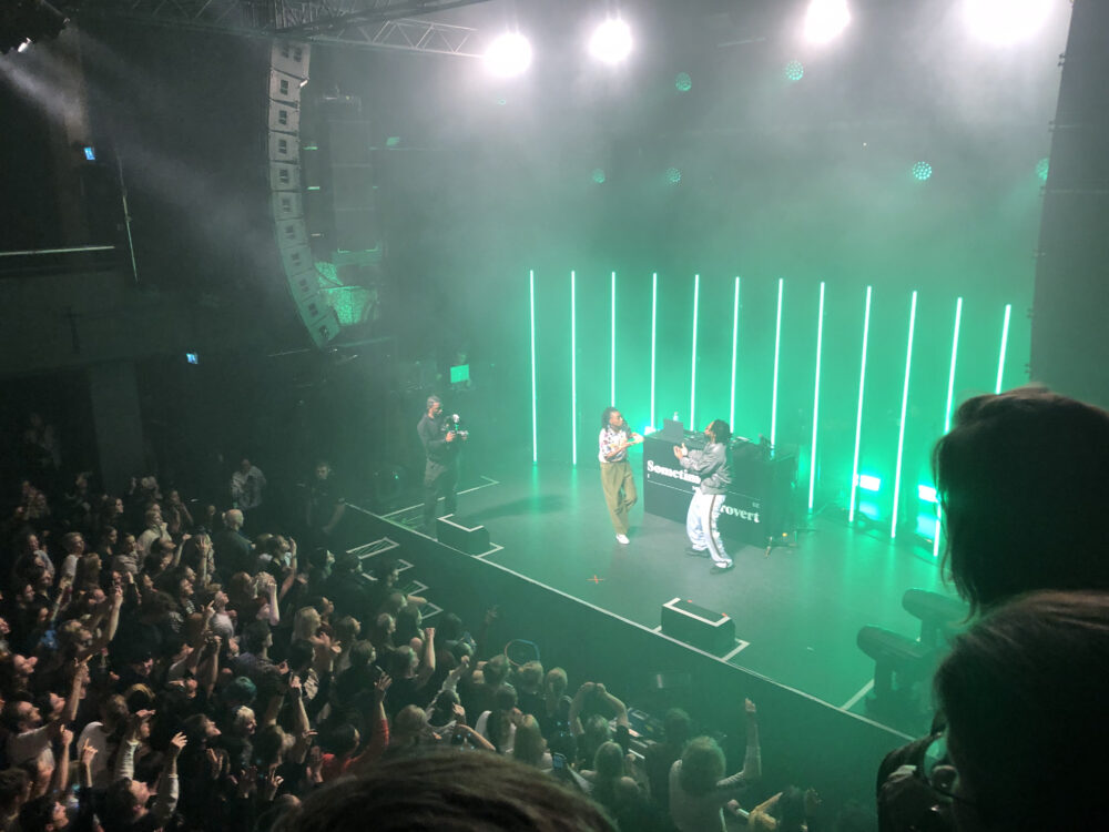 A photo, taken from a balcony, of a music venue. From this angle, you can see the crowd and the stage. On stage the artist Little Simz is dancing around the stage with her backing DJ.